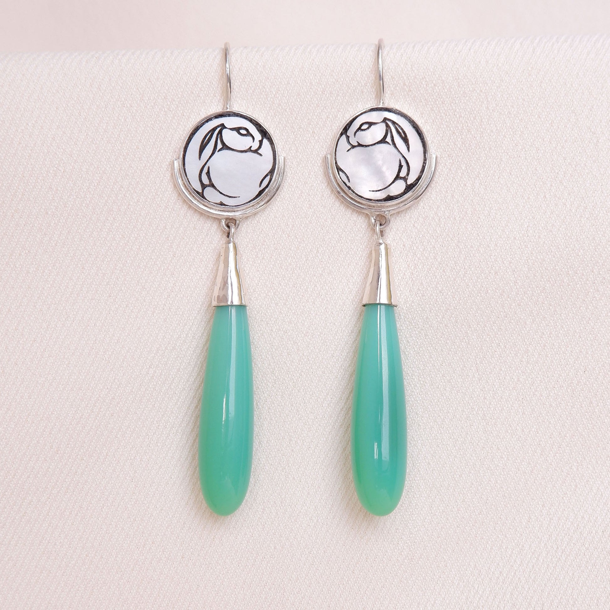 sterling silver earrings featuring mother of pearl discs, engraved with a blackened rabbit design, and adorned with long green smooth chrysoprase gemstone drop pendants. shown on a cream silk background