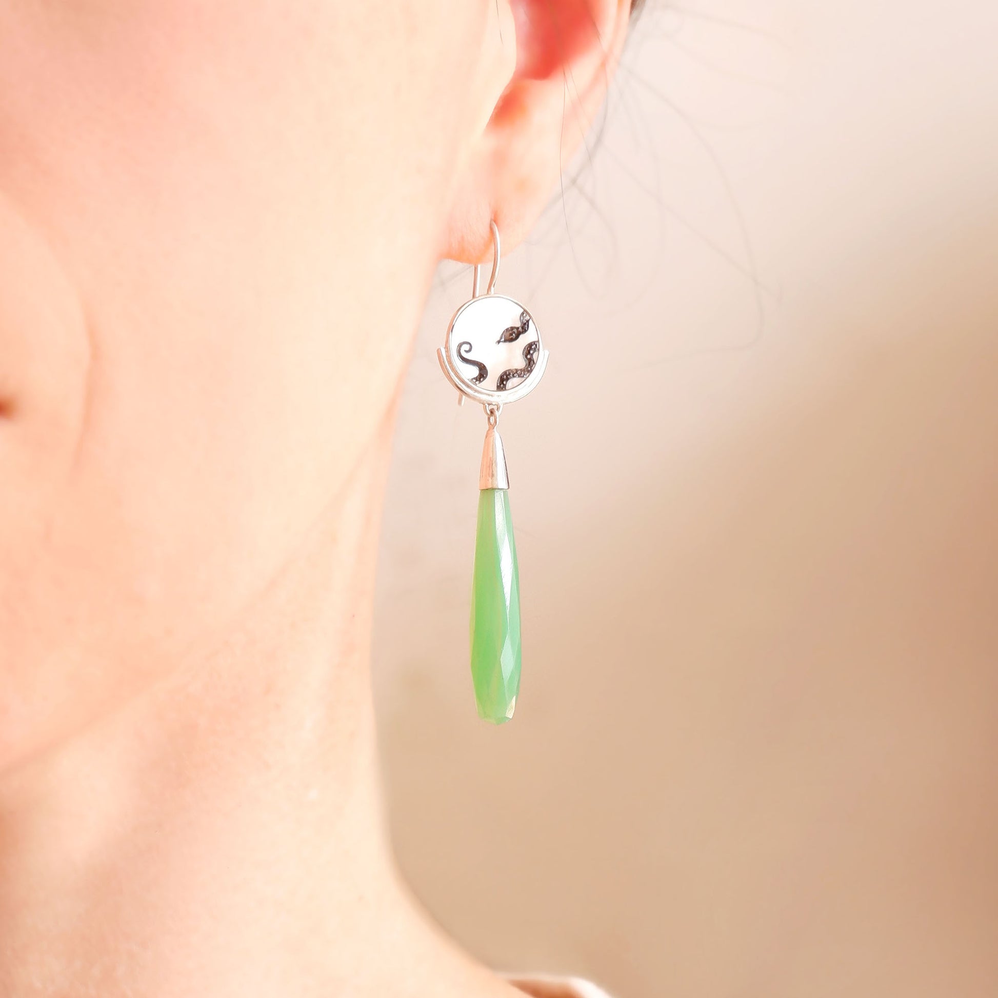 sterling silver earrings featuring mother of pearl discs, engraved with a blackened snake design, and adorned with long green faceted chrysoprase gemstone drop pendants. Shown on a model, cropped.