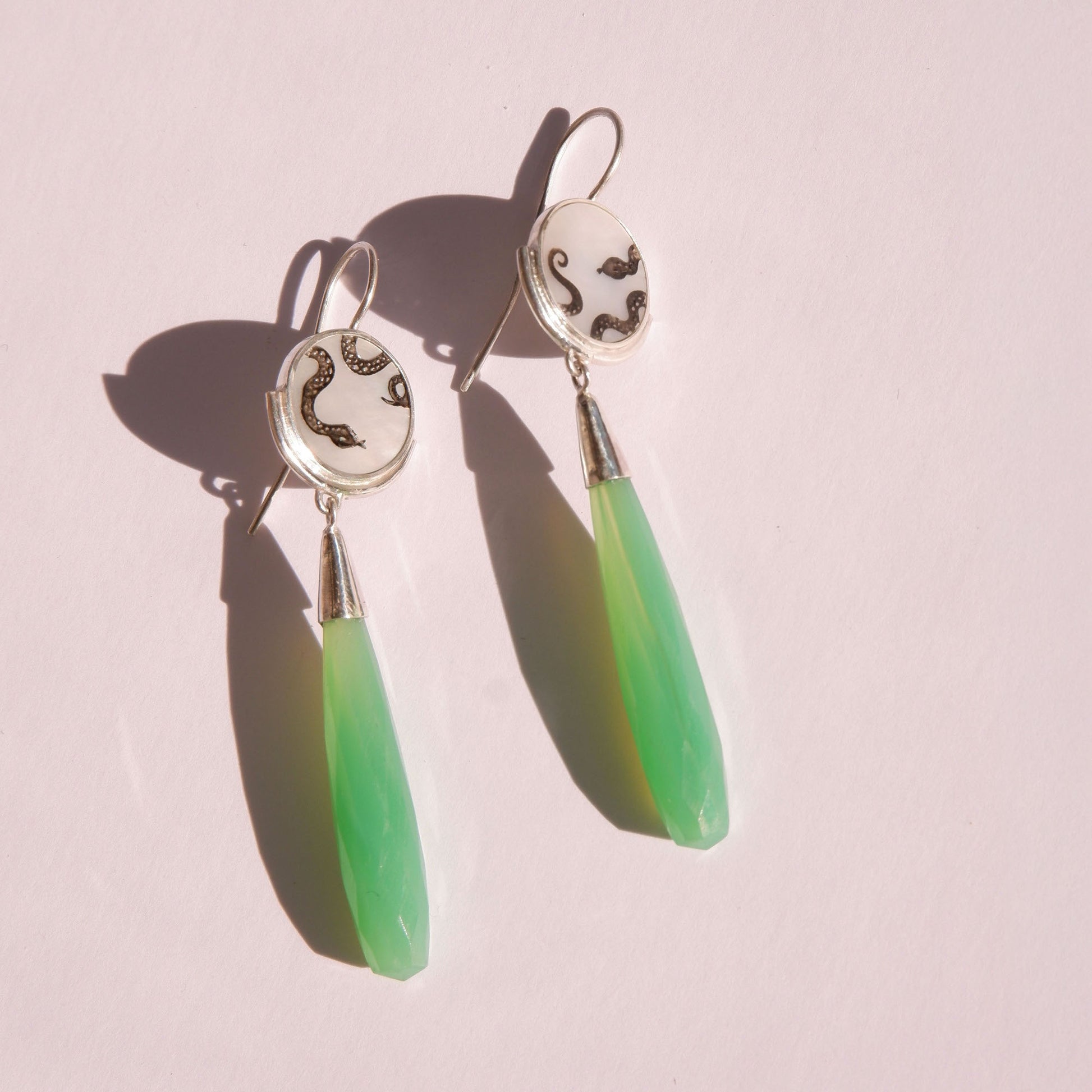 sterling silver earrings featuring mother of pearl discs, engraved with a blackened snake design, and adorned with long green faceted chrysoprase gemstone drop pendants. Shown laying in bright sunlight.
