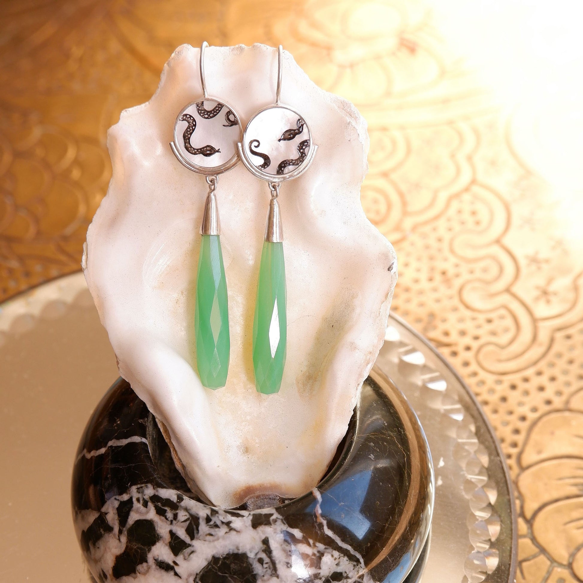 sterling silver earrings featuring mother of pearl discs, engraved with a blackened snake design, and adorned with long green faceted chrysoprase gemstone drop pendants. Shown hanging from an oyster shell in the sunshine.