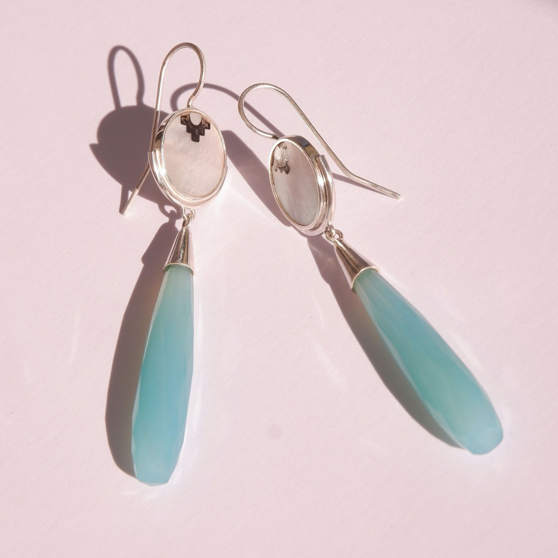 sterling silver earrings featuring mother of pearl discs, engraved with a blackened geometric steps design, and adorned with long blue faceted chalcedony gemstone drop pendants. Shown sitting flat, in bright sunlight