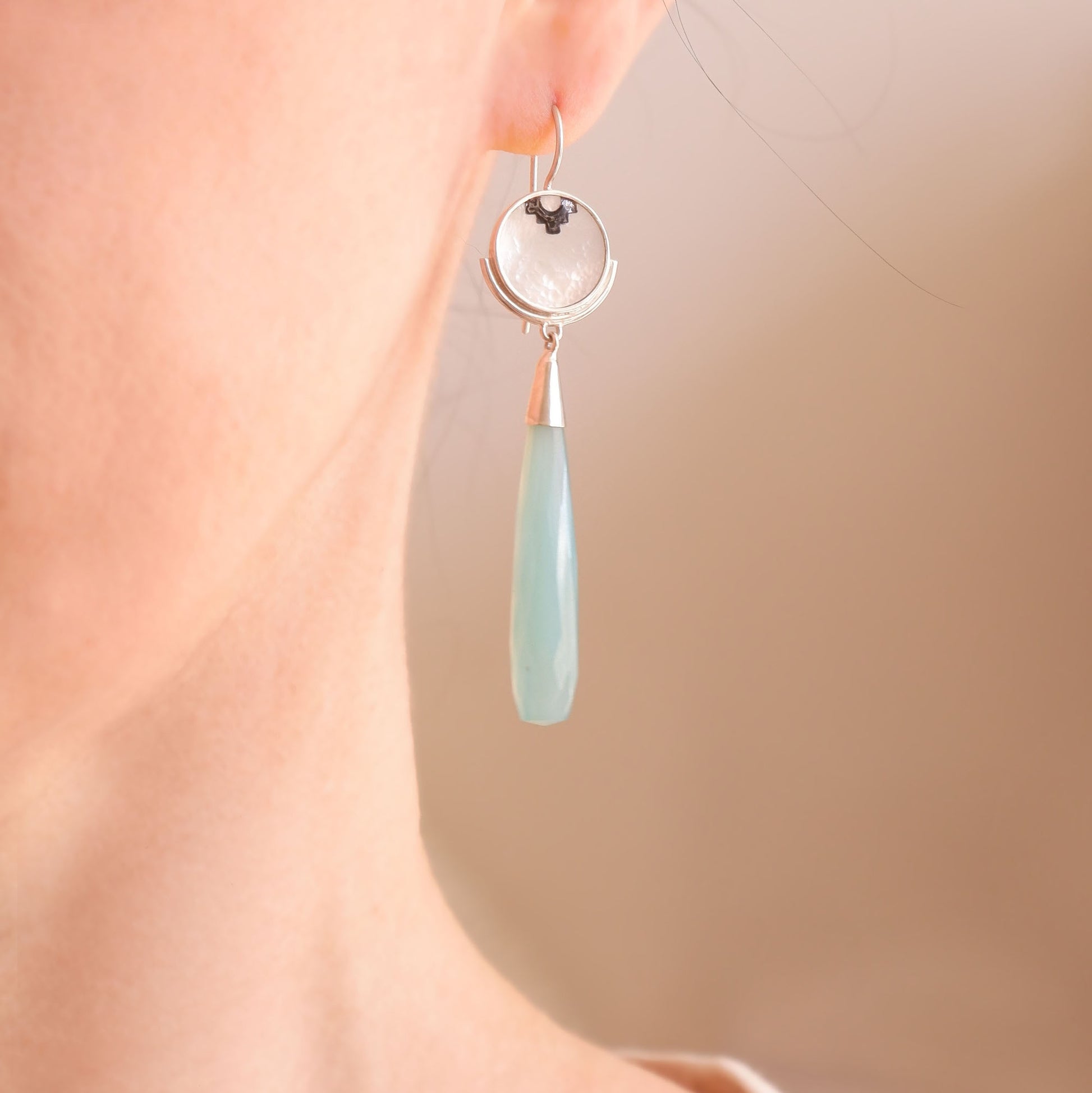 sterling silver earrings featuring mother of pearl discs, engraved with a blackened geometric steps design, and adorned with long blue faceted chalcedony gemstone drop pendants. Shown on model, close up
