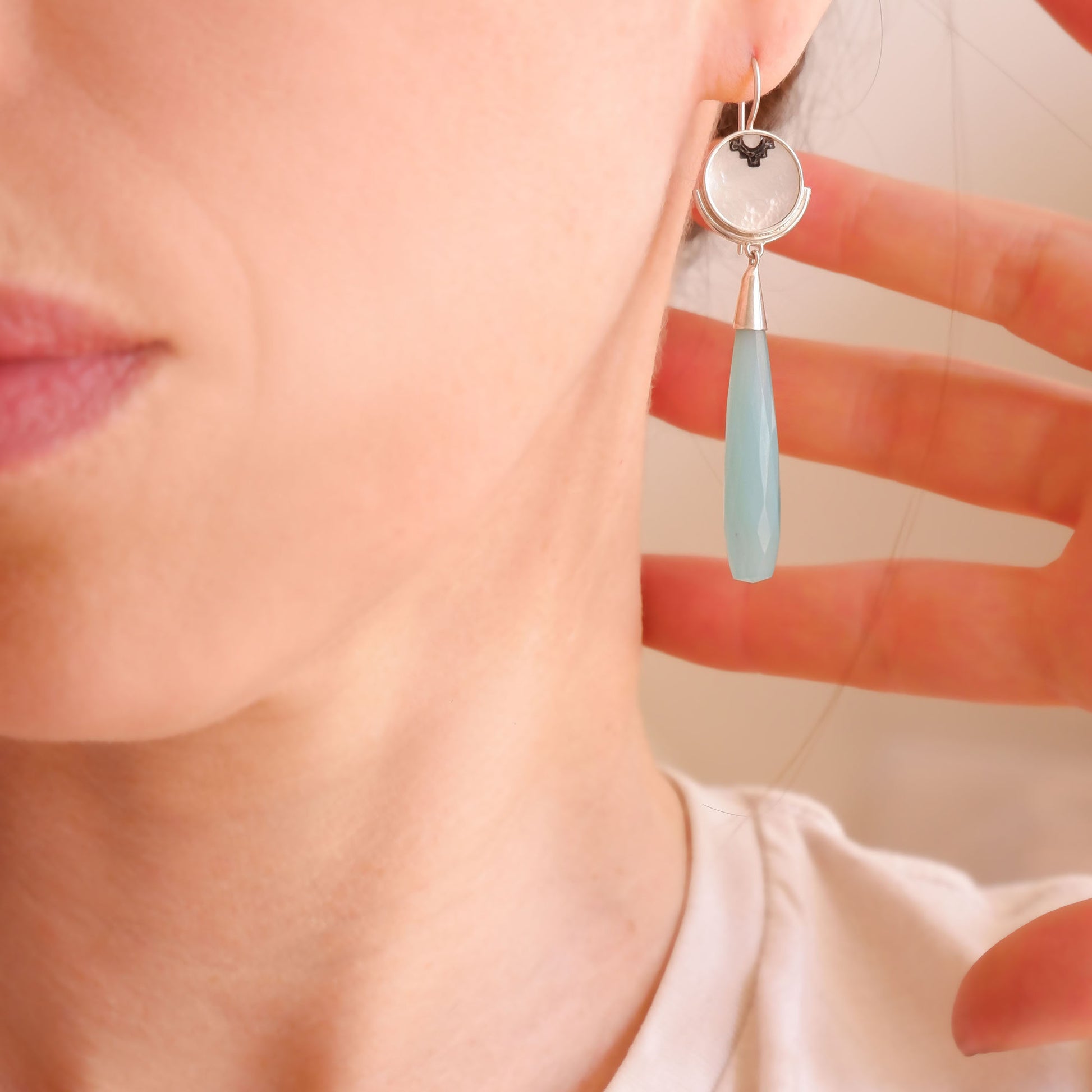 sterling silver earrings featuring mother of pearl discs, engraved with a blackened geometric steps design, and adorned with long blue faceted chalcedony gemstone drop pendants. Shown on model, close up