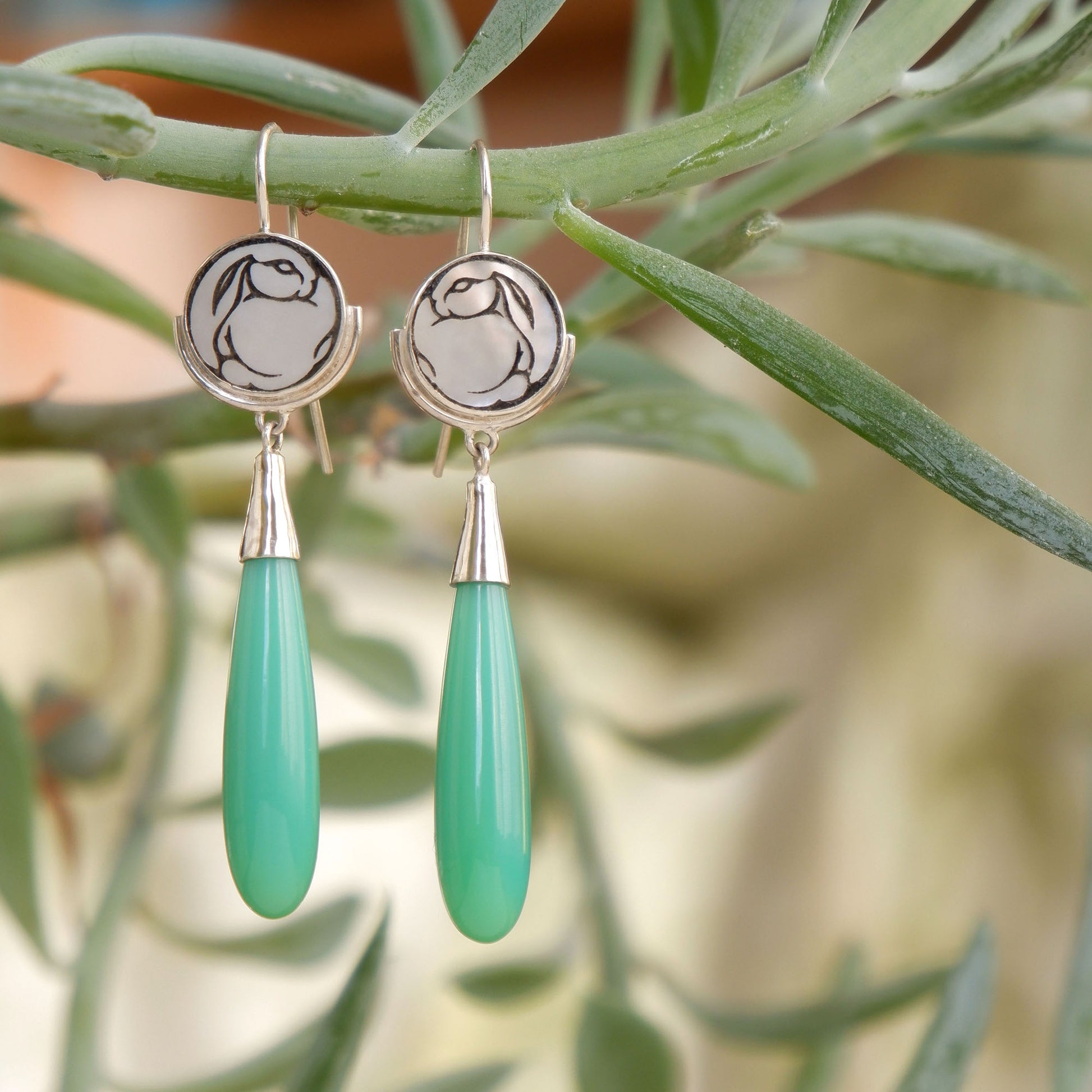 sterling silver earrings featuring mother of pearl discs, engraved with a blackened rabbit design, and adorned with long green smooth chrysoprase gemstone drop pendants. shown hanging from a plant