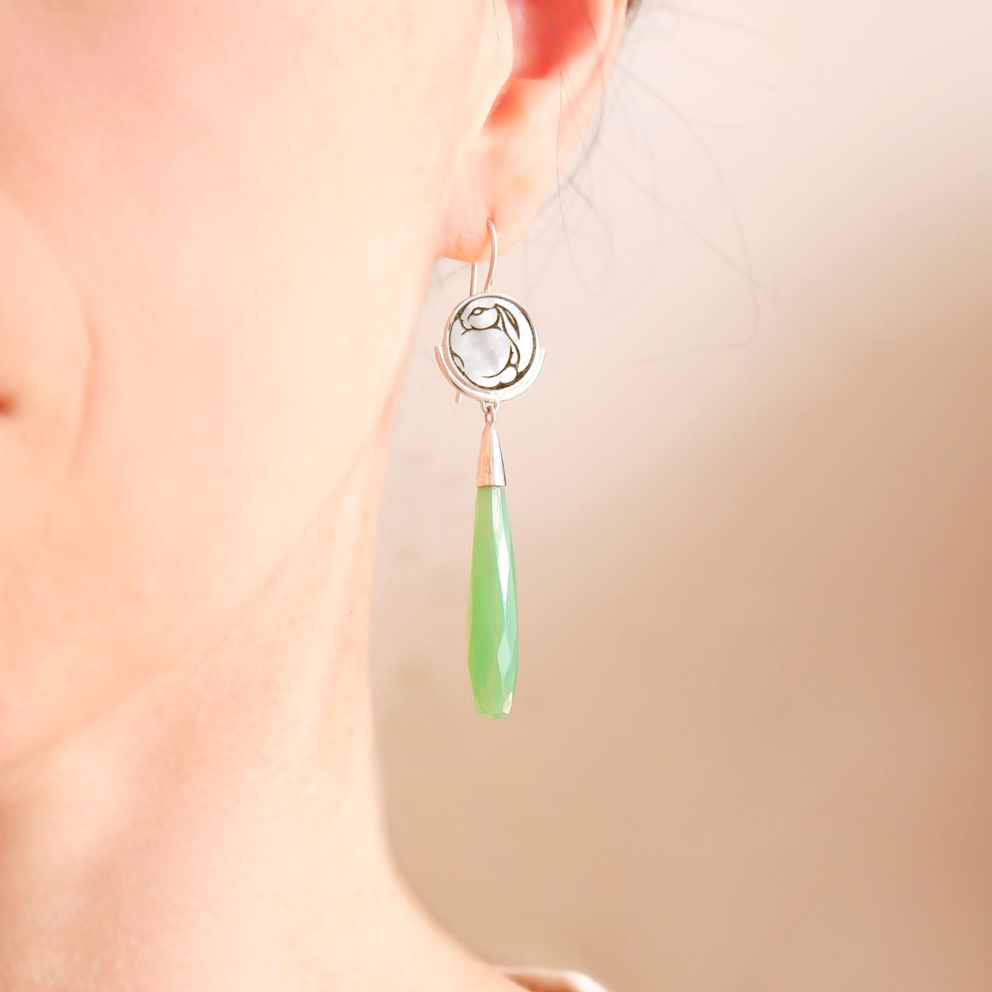 sterling silver earrings featuring mother of pearl discs, engraved with a blackened rabbit design, and adorned with long green smooth chrysoprase gemstone drop pendants. shown on model
