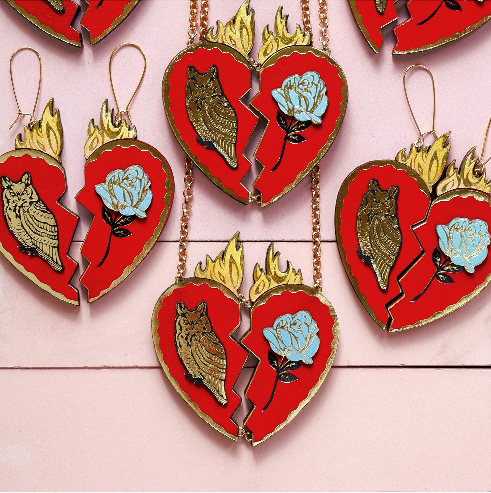 red leather broken heart pendants & earrings, with golden owls, flames & blue roses,  laid out flat on pink background