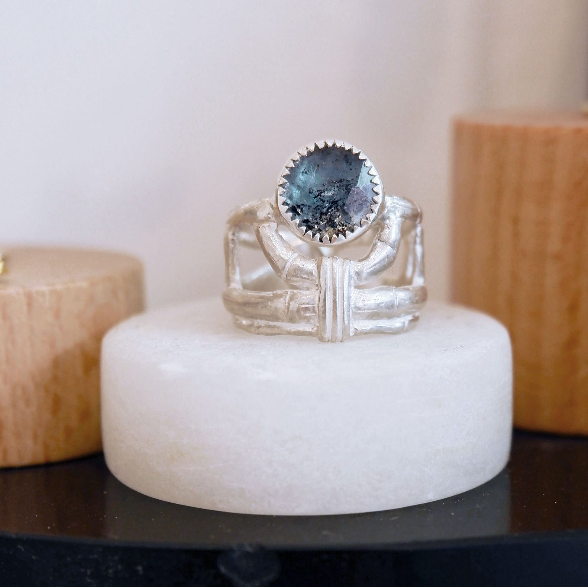 structural open-work ring in a bamboo style, featuring a faceted round teal blue Kyanite gemstone in a serrated setting. Sterling Silver. Shown sitting on a small plinth