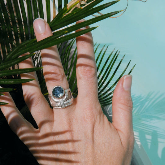 structural open-work ring in a bamboo style, featuring a faceted round teal blue Kyanite gemstone in a serrated setting. Sterling Silver. Shown sitting on a hand for scale, with palm leaf & a blue pool