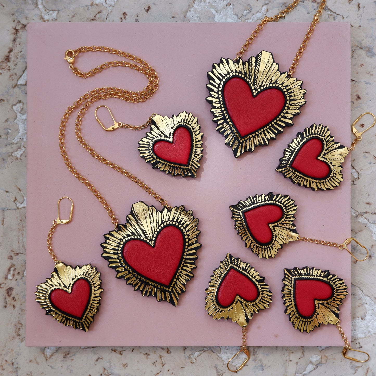 display of red leather sacred heart pendant necklaces, on gold belcher chain with matching red leather sacred heart earrings, on pink background