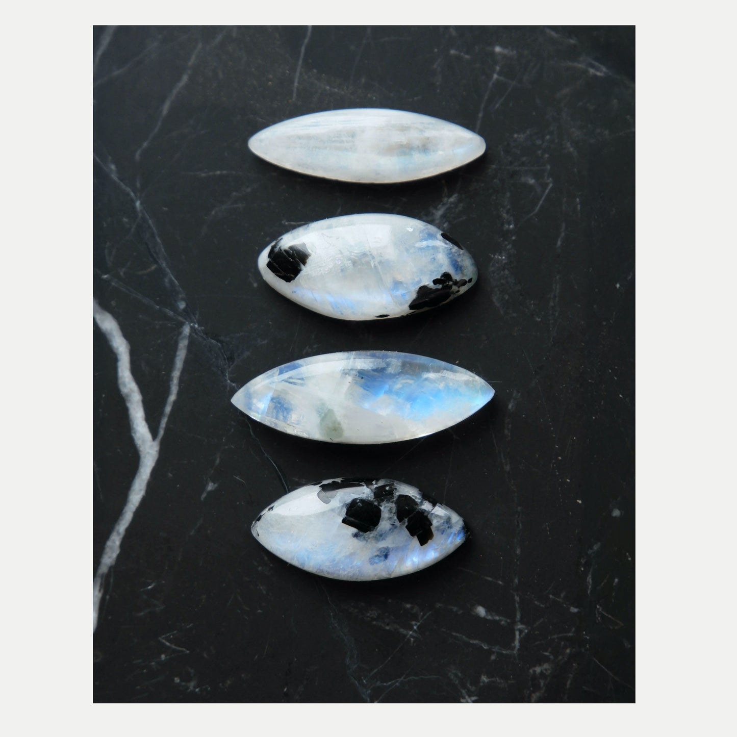 four unique Marquise-Cut faceted Moonstone Gemstones, two are white/ rainbow moonstone, two have distinctive Black Tourmaline inclusions. Shown on black marble tile.
