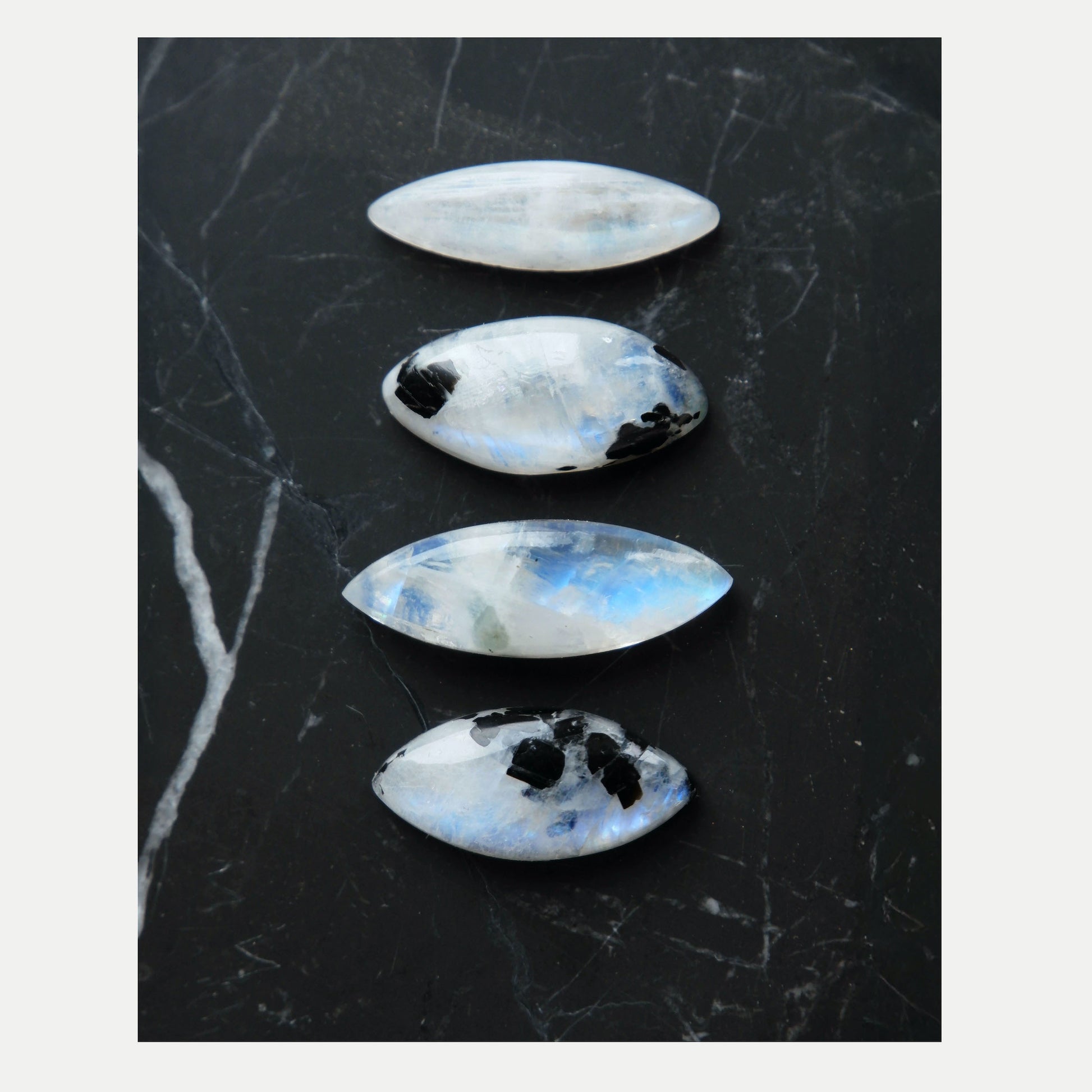 four unique Marquise-Cut faceted Moonstone Gemstones, two are white/ rainbow moonstone, two have distinctive Black Tourmaline inclusions. Shown on black marble tile.