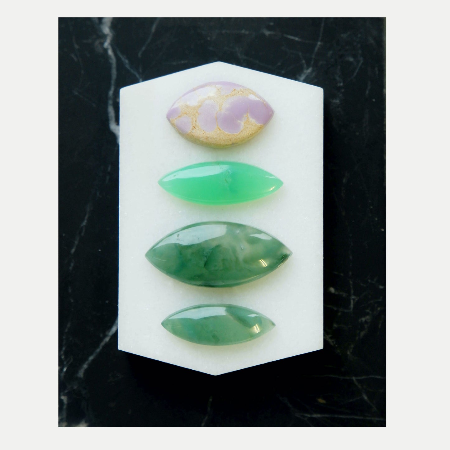 Four different Marquise-Cut  Gemstone Cabochons, Shown on black marble tile. Marbled Lilac & gold Phosphosiderite, bright Green Chrysoprase, Dark swirling Green Chalcedony