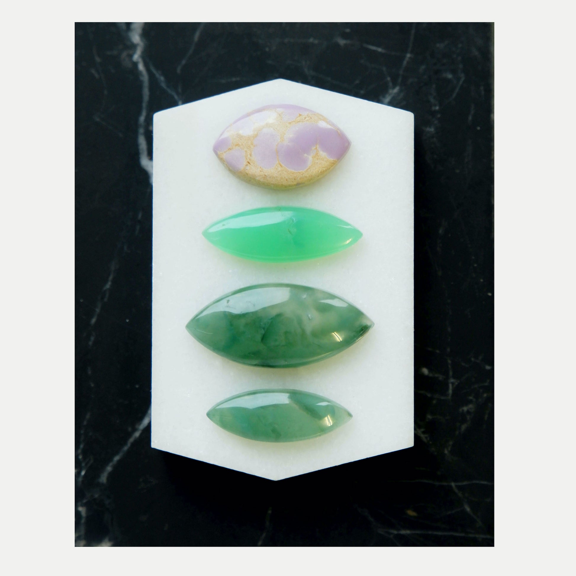 Four different Marquise-Cut  Gemstone Cabochons, Shown on black marble tile. Marbled Lilac & gold Phosphosiderite, bright Green Chrysoprase, Dark swirling Green Chalcedony