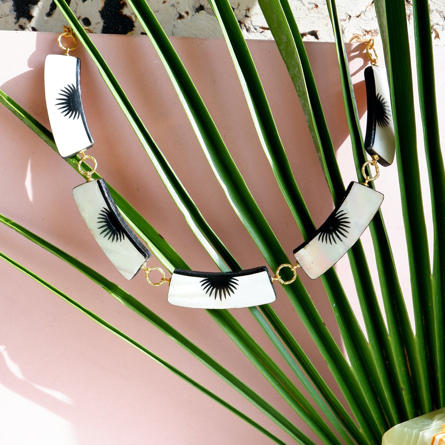 graphic black & white palm design mother of pearl necklace with 5 panels, on golden long-link chain, lifestyle shot with palm leaf