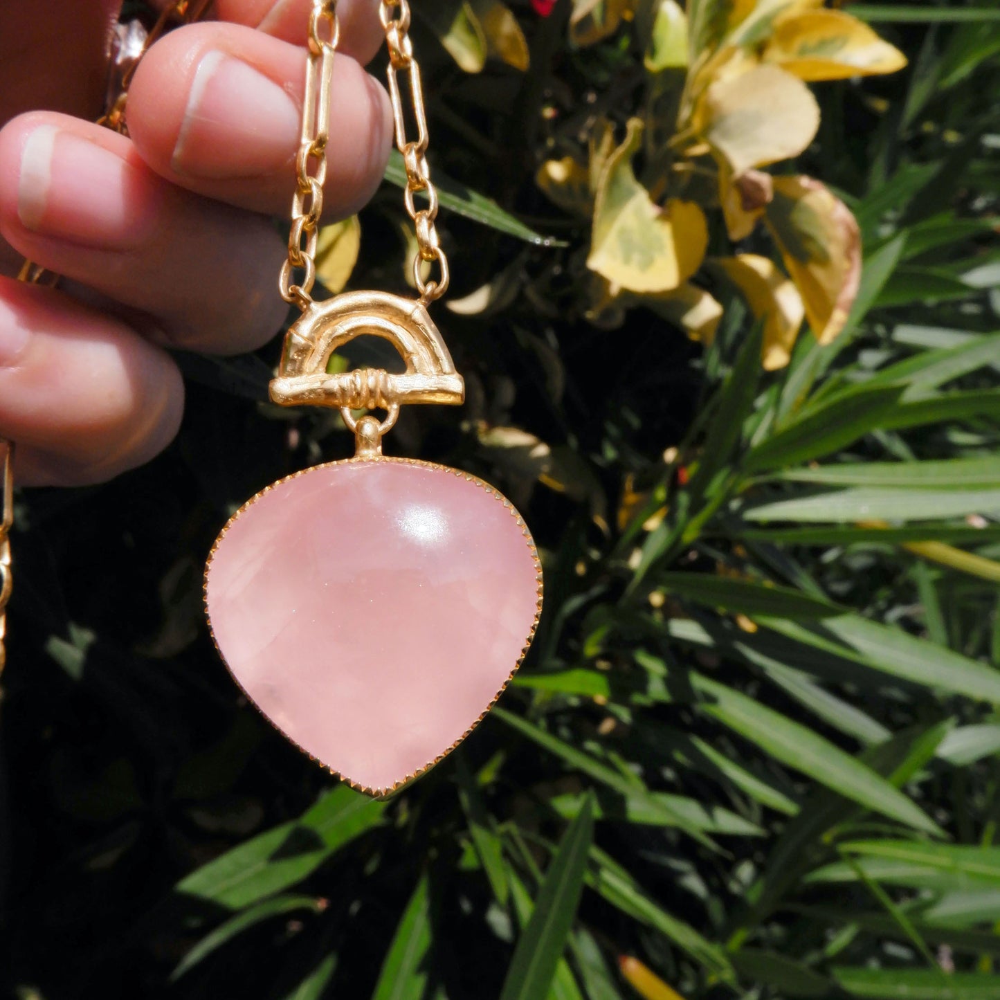 ose pink quartz heart-shaped pendant with golden bamboo arch detail, on a gold chain, against a tropical foliage background