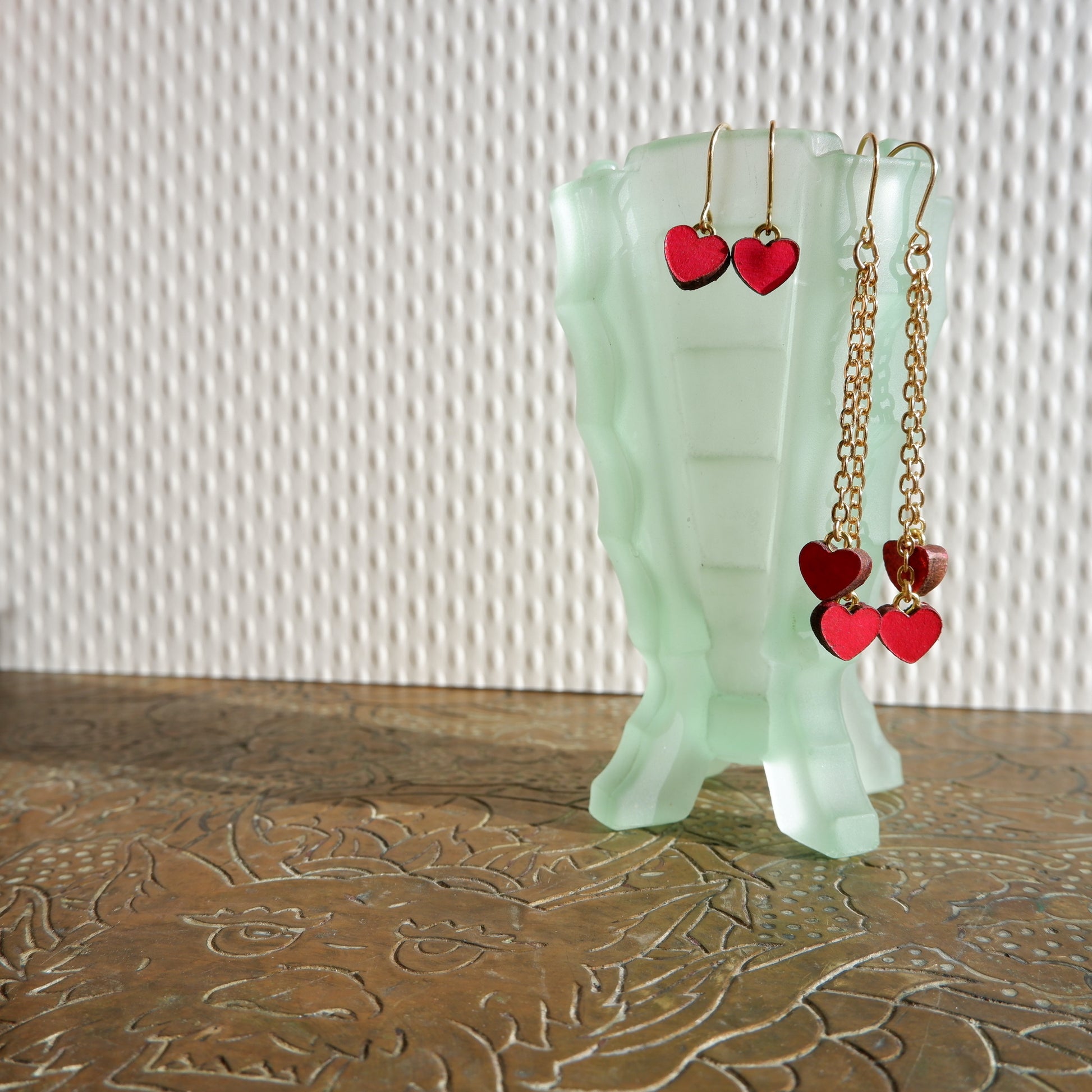 two pairs of small red leather hearts earrings hanging from a pale green vintage glass vase