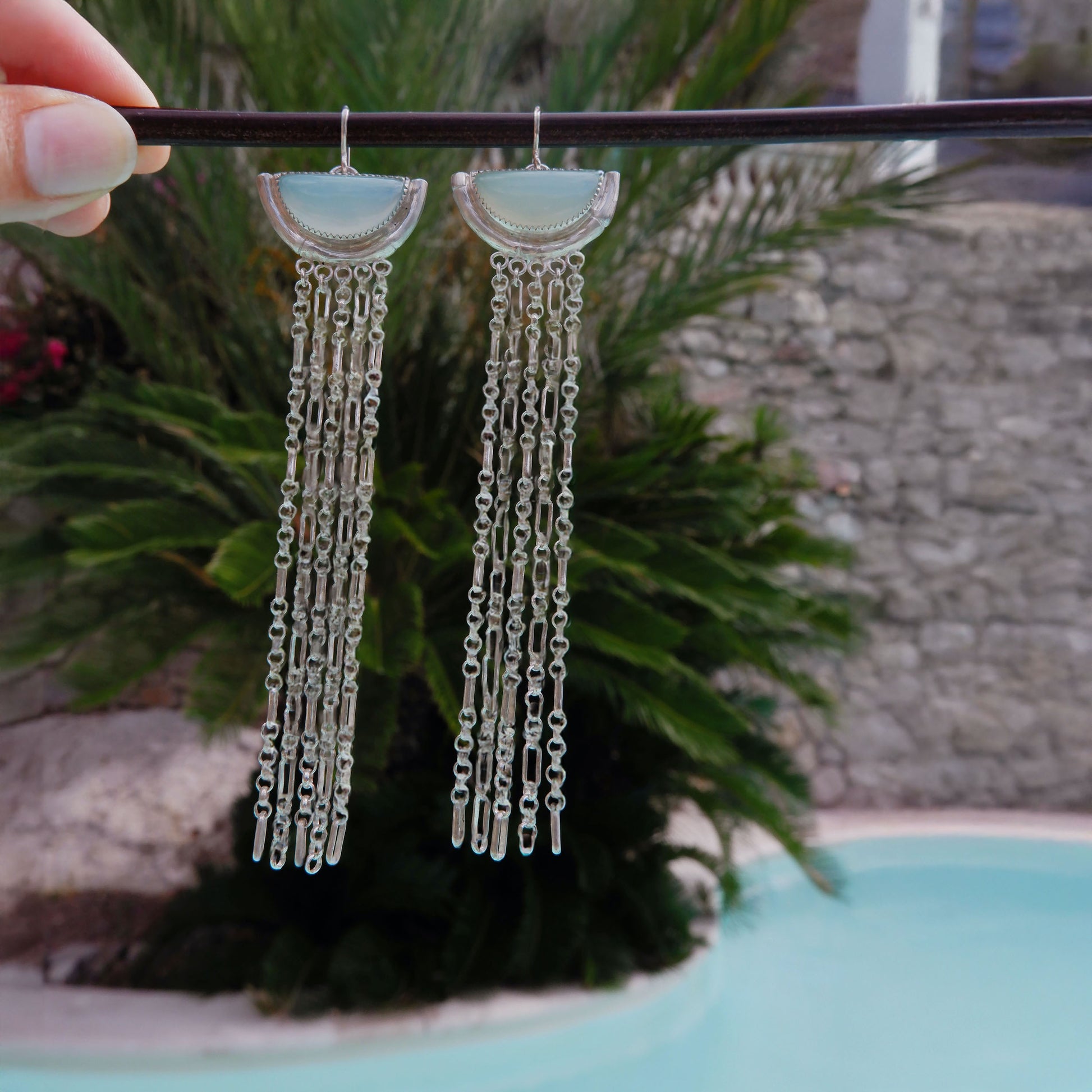 dramatic long chain fringe chandelier tassel earrings with half moon shaped semi precious aqua blue chalcedony gemstones,  cupped by solid silver bamboo hammock, with simple silver wire hooks. Shot agains palm tree & aqua blue swimming pool.