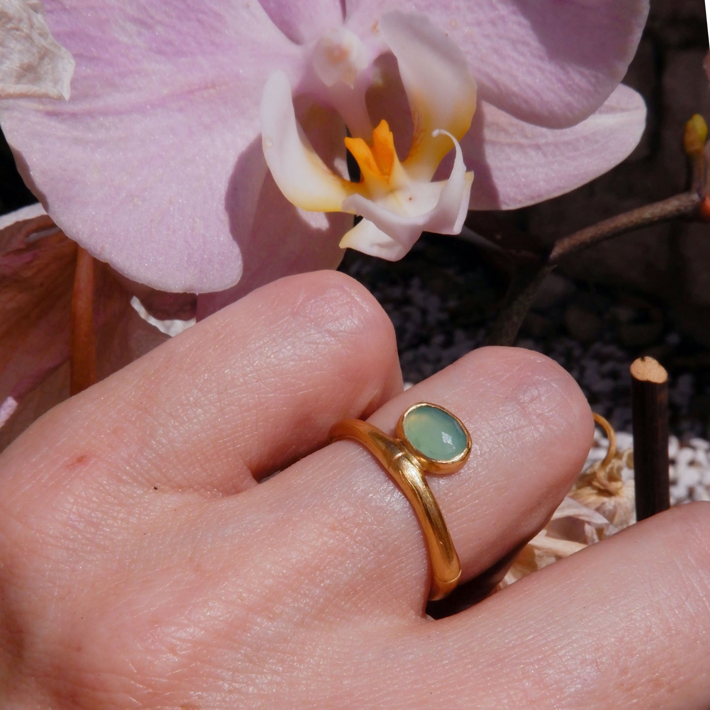 gold bamboo style ring with a small faceted oval pale apple green Chrysoprase gem stone, on a hand in strong sunlight, with a pink orchid