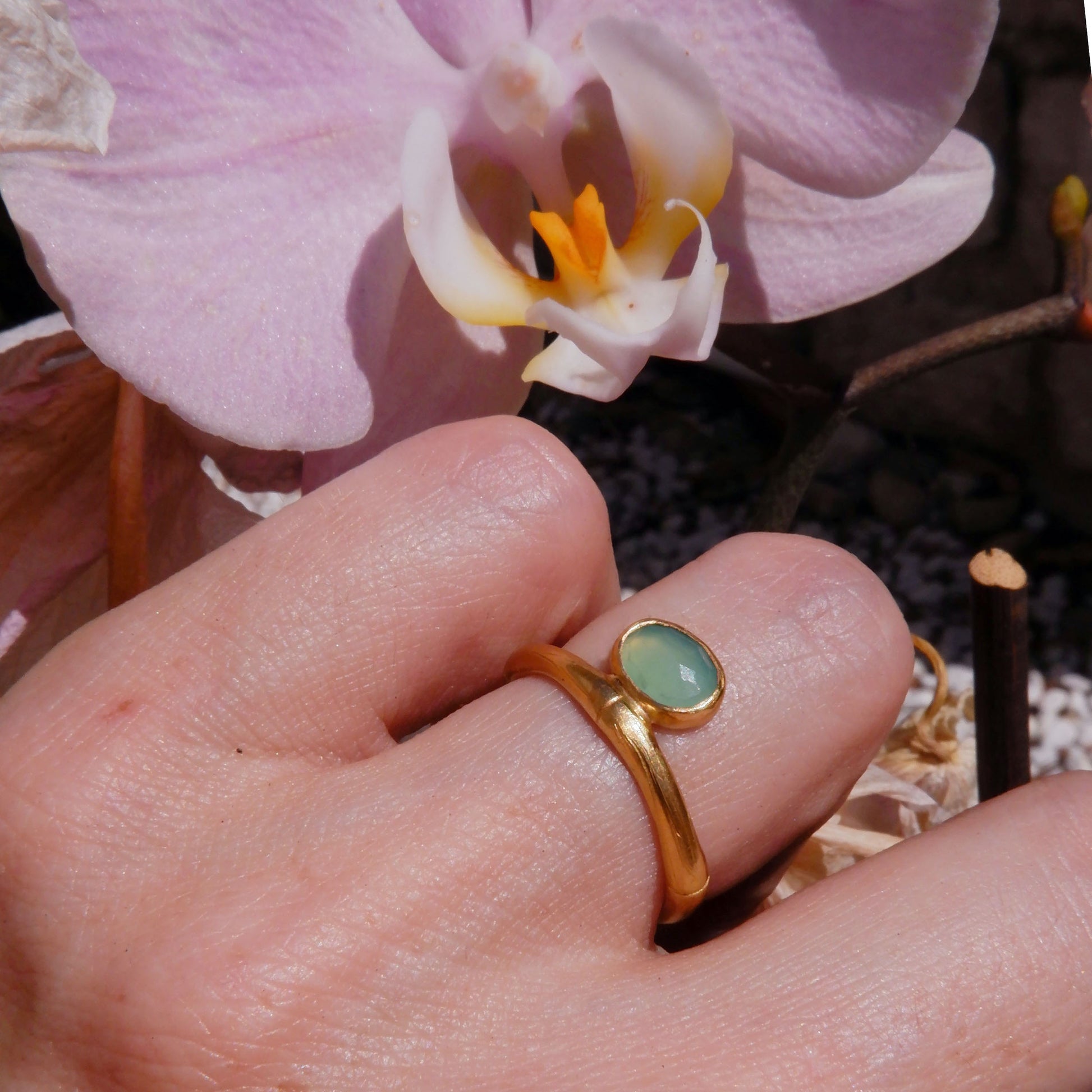 gold bamboo style ring with a small faceted oval pale apple green Chrysoprase gem stone, on a hand in strong sunlight, with a pink orchid