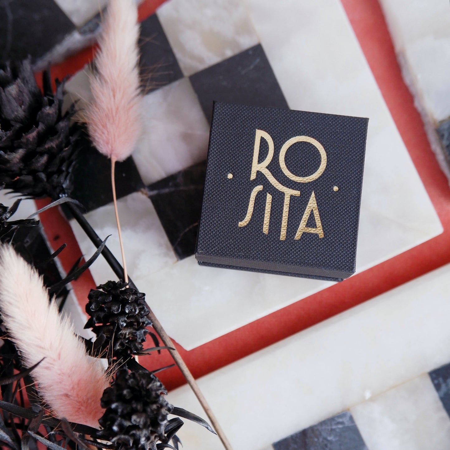 small square Black ring box with 'ROSITA' branding printed in gold. background of black & white chess board, pink velvet, black & pink dried flowers