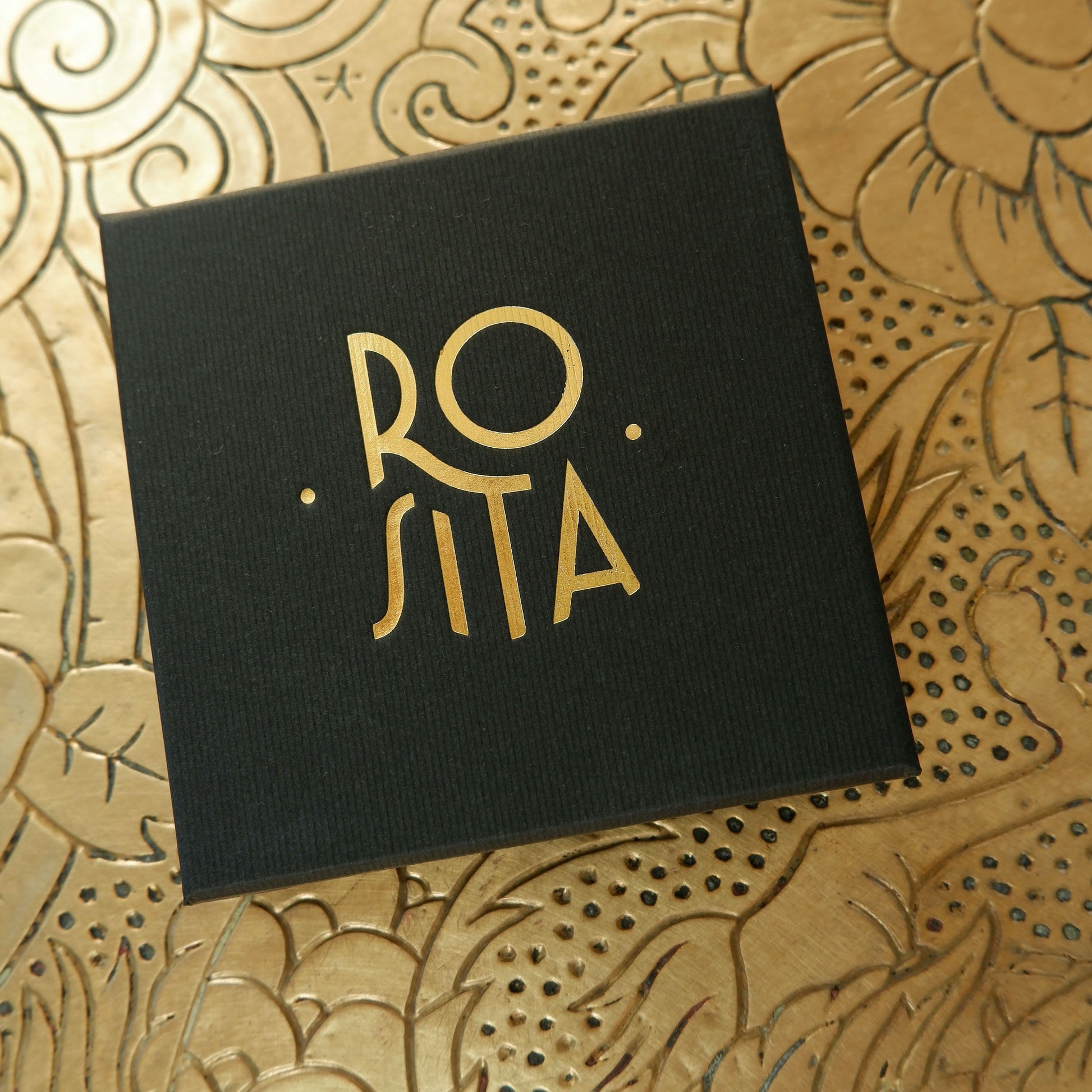 Black finely ribbed cardboard jewellery box, with ROSITA  logo branding in Gold foil, on gold patterned background