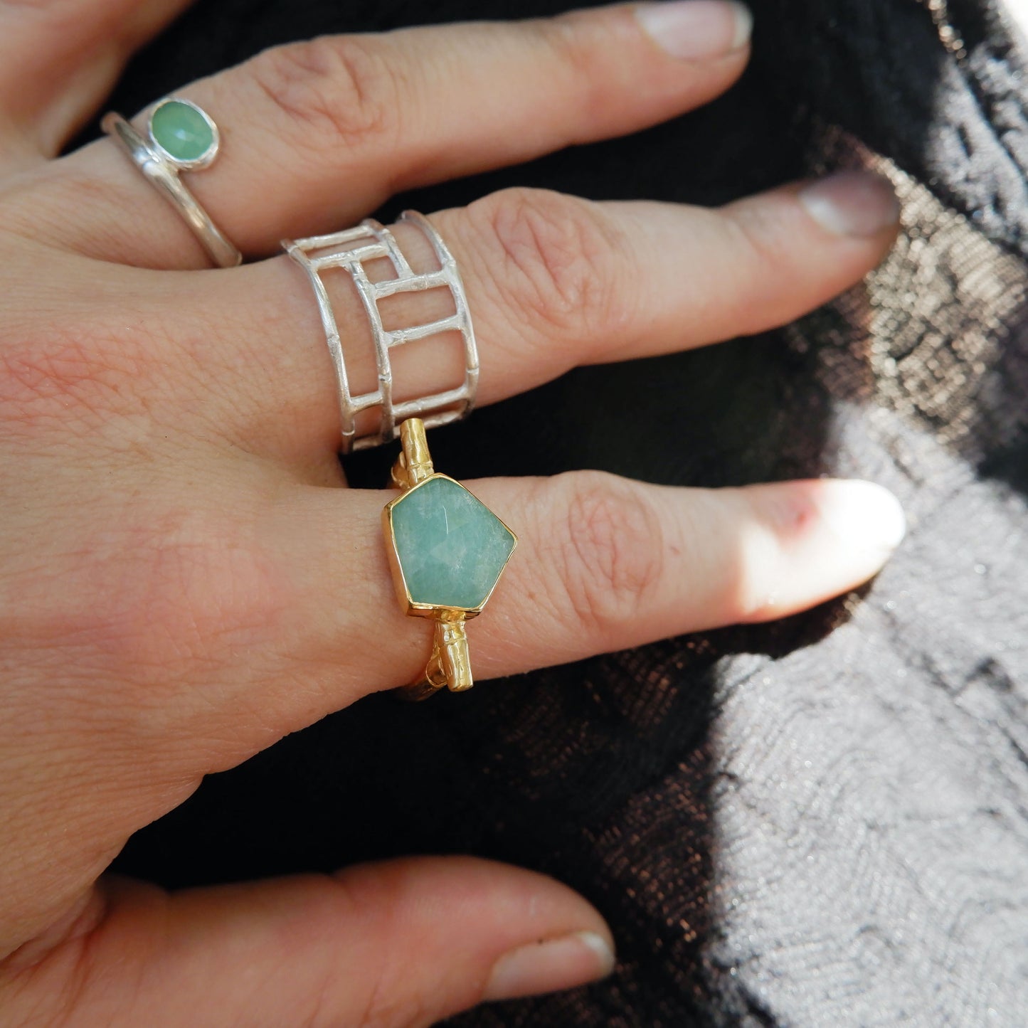Gold flat top bamboo style ring with a an irregular angular geometric Amazonite gem stone, on a hand with other bamboo style rings in sterling silver, against black cloth