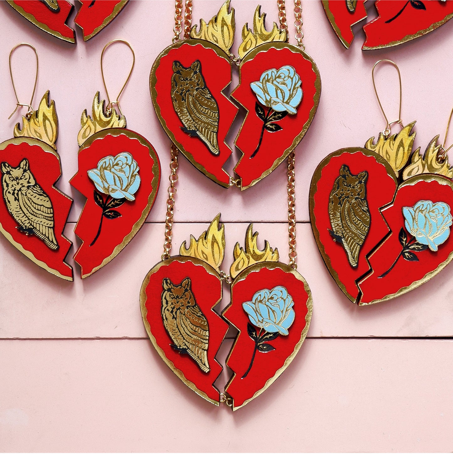  red leather broken heart pendants & earrings, with golden owls, flames & blue roses,  laid out flat on pink background