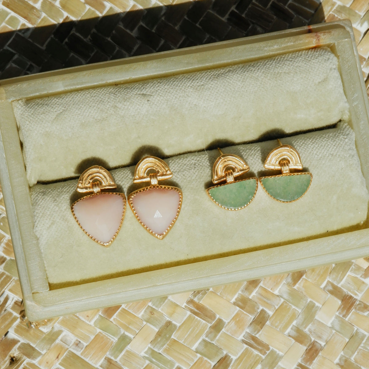Two pairs of Gold Vermeil stud drop earrings , featuring triple arches of bamboo, paired with hanging gemstone charm pendants, set with serrated gold teeth. 
One pair has Trillion Shaped Pink Opal, the other Half moon shapedGreen Aventurine. Shown in bright sunlight in a pale green velvet box