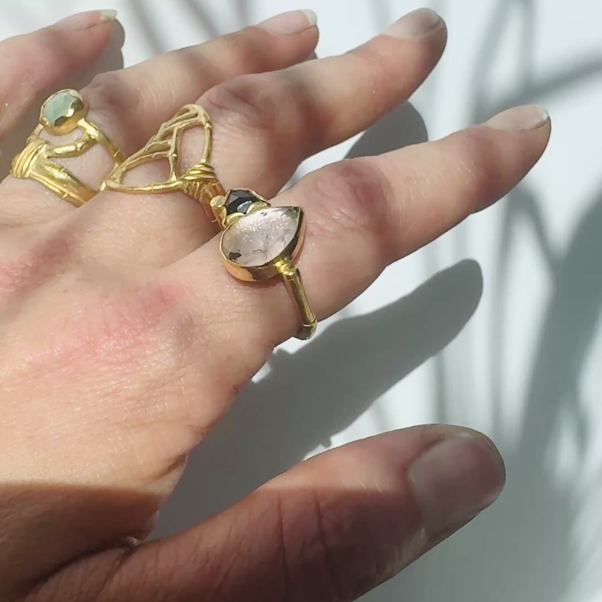 video showing Gold ring with geometric open-work Bamboo Circle screen design, on a white background, another openwork bamboo style ring with a round aquamarine gemstone, an da flat-top golden bamboo ring with a pear shaped rose quarts and black onyx trillion cut gemstone, on a hand.
