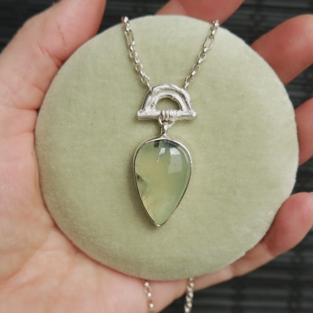 video showing Pendant with light green Prehnite pear shaped gemstone hanging from silver Bamboo Arch, and silver chain.  shown on pale green velvet