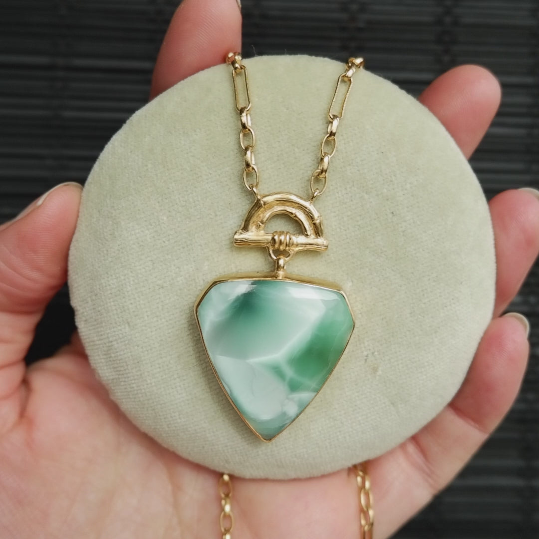 video showing Pendant with large shield shaped green hemimorphite gemstone hanging from Gold Bamboo Arch, a long doubled gold chain.  shown on pale green velvet