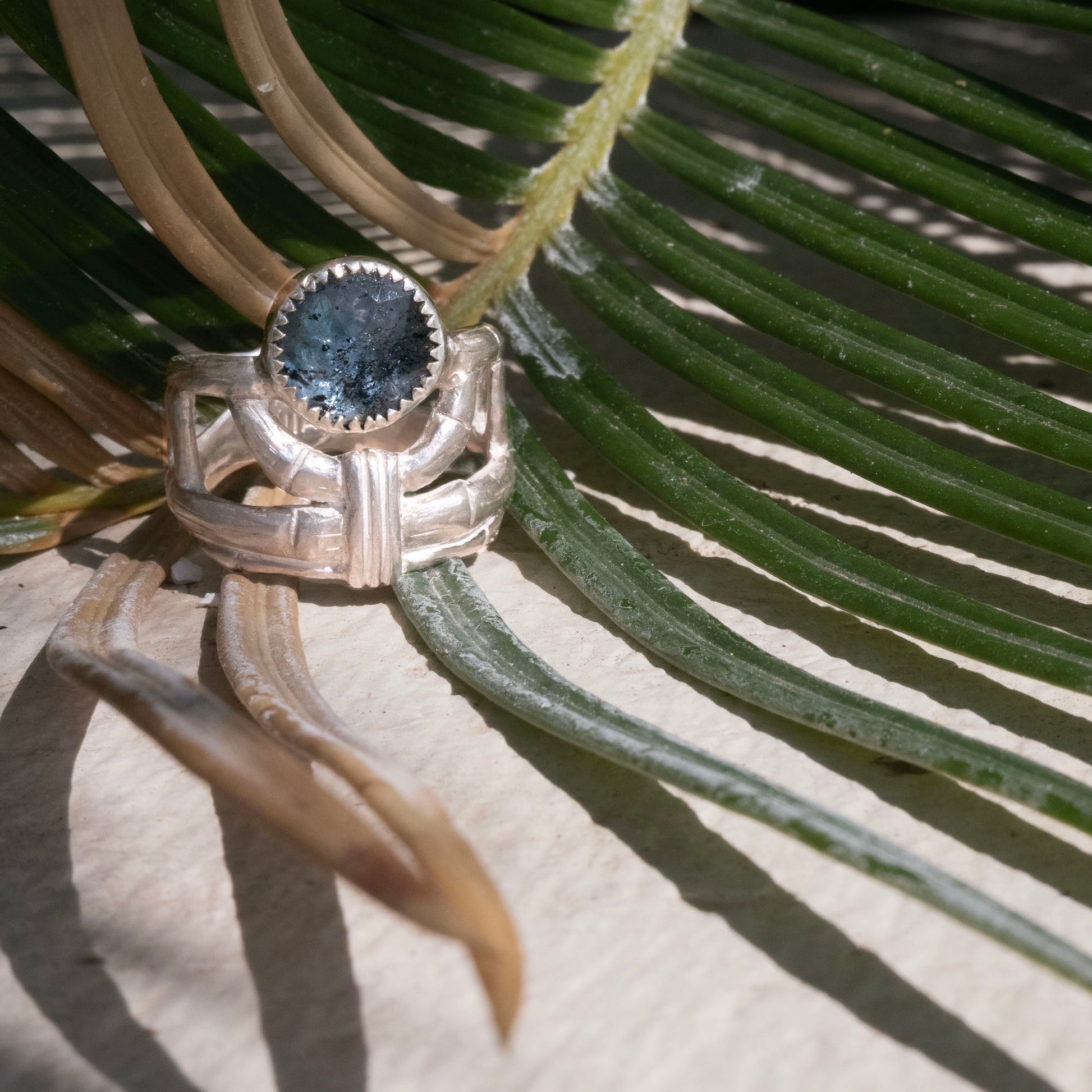structural open-work ring in a bamboo style, featuring a faceted round teal blue Kyanite gemstone in a serrated setting. Sterling Silver. Shown sitting on a h palm leaf 