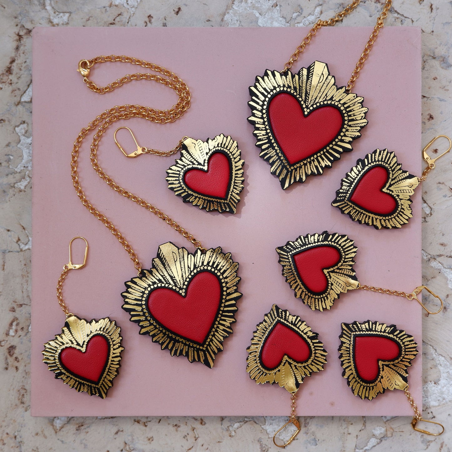 display of  red leather sacred heart pendant necklaces, on gold belcher chain with matching red leather sacred heart earrings, on pink background